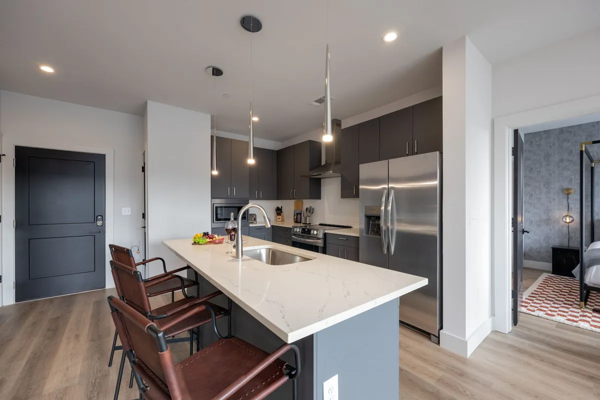 This image features a modern kitchen with a spacious island, stainless steel appliances, and sleek cabinetry. The island includes bar stools for casual dining or social interaction during meal prep. The kitchen is brightly lit and designed for both functionality and entertaining.