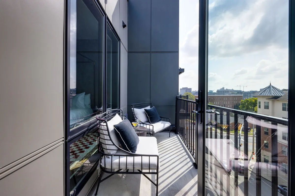 A serene balcony scene with comfortable seating and a small table, looking out over the city through glass doors. The setting is perfect for enjoying morning coffee or evening drinks while taking in the views and fresh air.