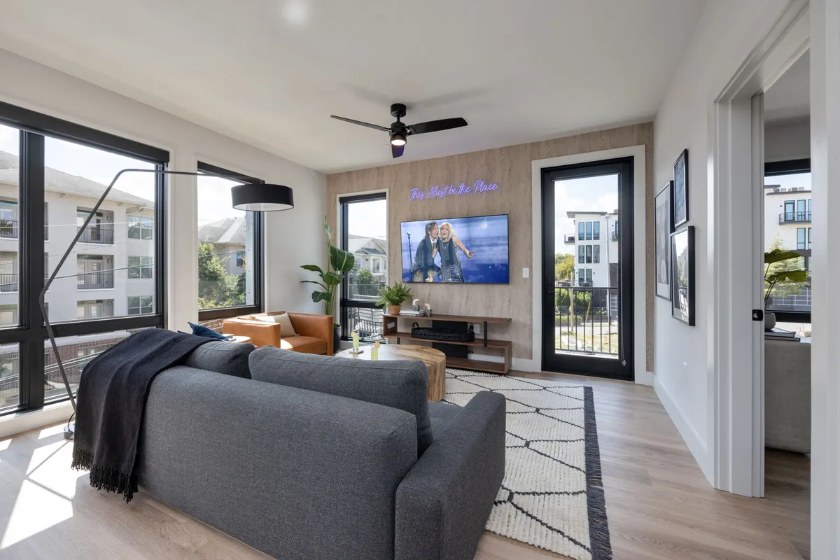A stylish living room featuring a plush sofa, coffee table, and a Smart TV set in front of large windows that let in abundant natural light. The space is designed for relaxing, watching movies, or entertaining guests, embodying comfort and style.
