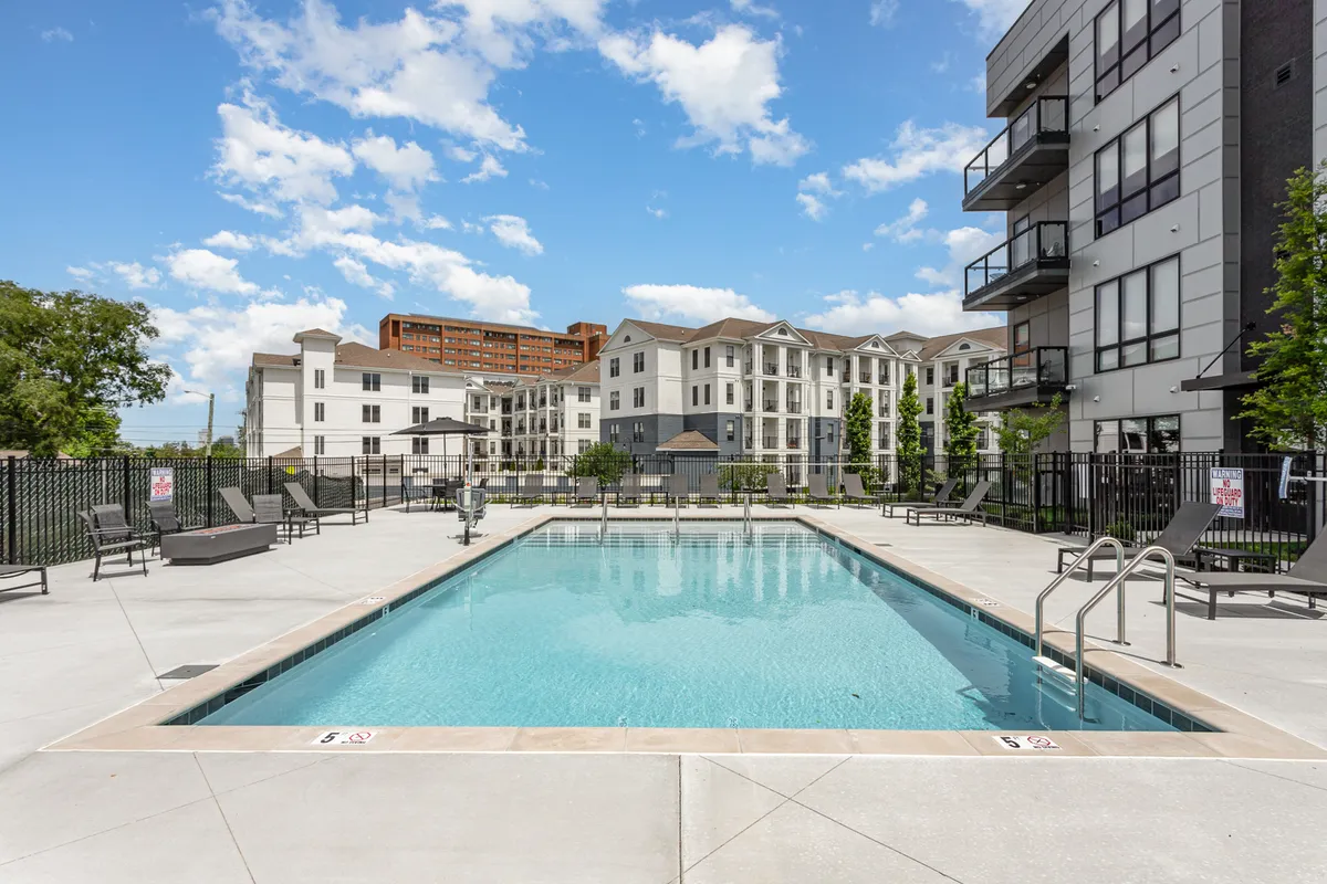 This exterior image captures a vibrant swimming pool area surrounded by modern apartment buildings and lush greenery. Lounge chairs are arranged poolside, creating an inviting atmosphere for sunbathing, swimming, and socializing in a resort-like setting.