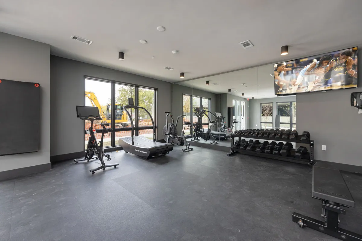 A brightly-lit, well-equipped fitness center with a treadmill, weights, an exercise bike, and expansive windows bringing in natural light. The space is inviting, energetic, and ideal for morning workouts or group fitness sessions.