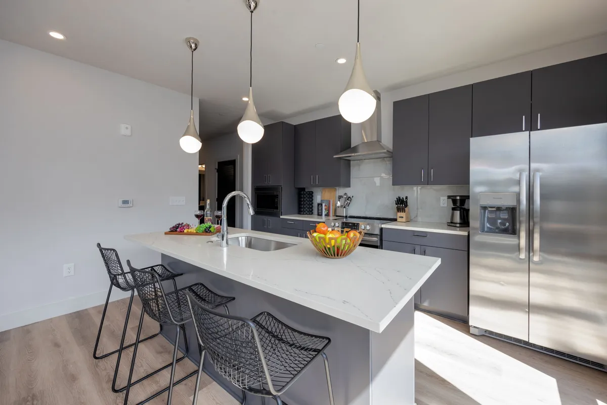 A friendly, functional kitchen scene centered on an island countertop. Hanging light fixtures and a bowl of fruit emphasize both practicality and warmth, inviting guests to cook, entertain, or enjoy family meals together.