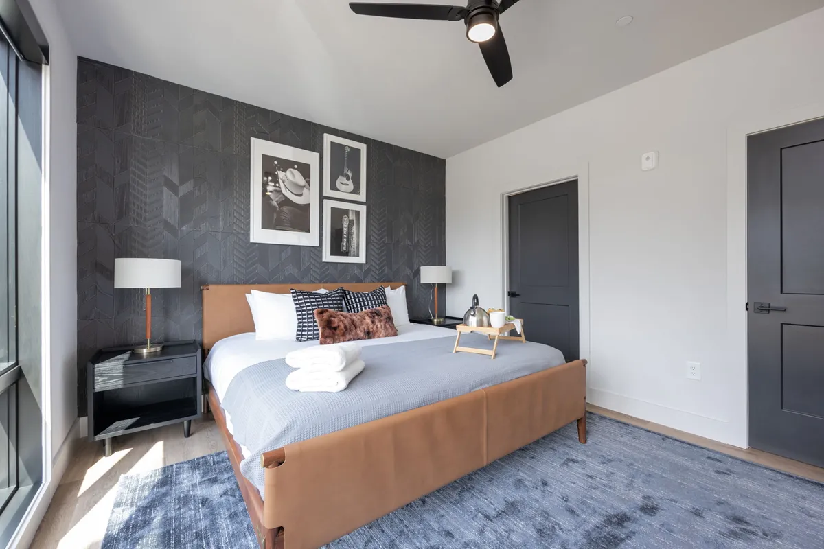 A king-size bed with a leather frame is centered in the bedroom, complemented by musical instrument-themed wall art, stylish bedside lamps, and decorative pillows. Morning sunlight streams in through the windows, creating a warm and inviting atmosphere ideal for relaxation or reading.