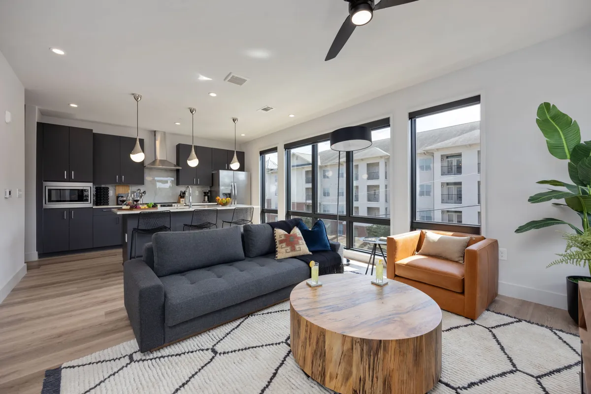 A bright, open living area with a plush sofa set near large, floor-to-ceiling windows. The wooden coffee table and kitchen area are visible, with potted plants adding a touch of greenery. Sunlight streams in, creating an inviting and comfortable atmosphere perfect for socializing, relaxing, or entertaining.