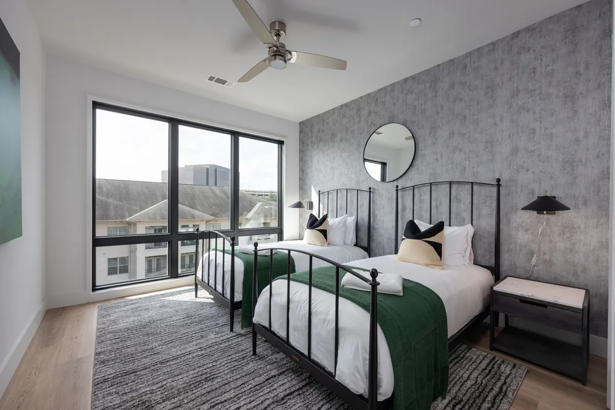 Two twin beds with black metal frames are positioned beside large windows, allowing natural light to flood the room. Modern round mirror, green throw blankets, and cozy pillows add a touch of comfort and style, making the space perfect for kids or friends.